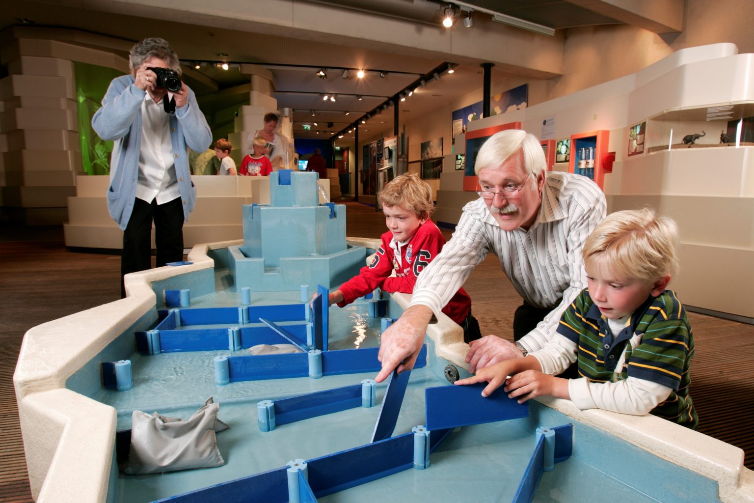 Eine Familie spielt an einem Wasserexperiment, das aus blauen Bahnen und Brettern besteht.