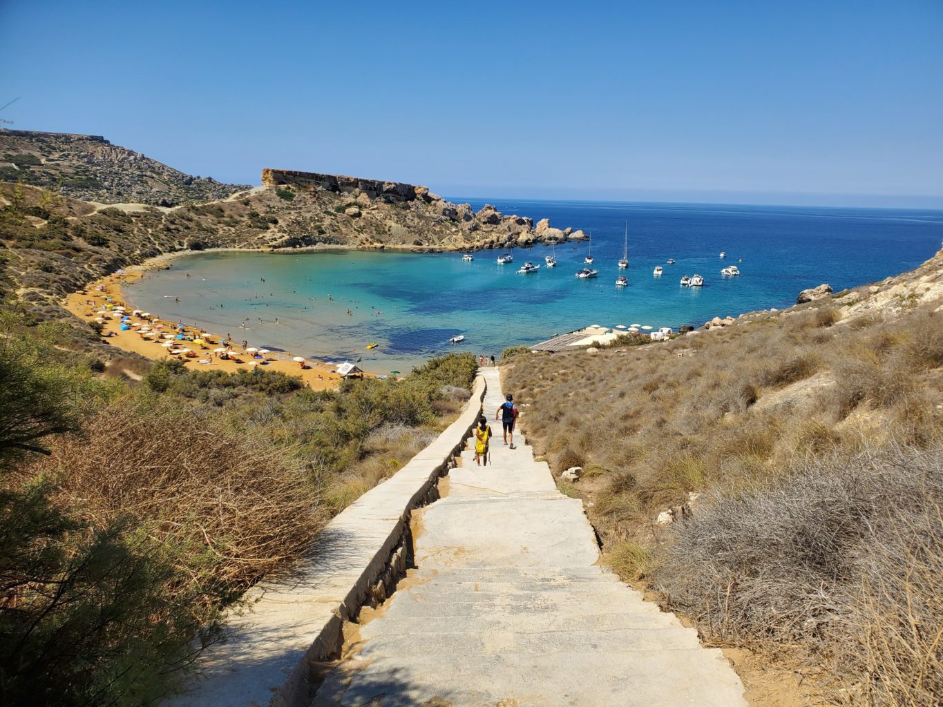 Eine lange Steintreppe führt in eine türkise Bucht, wo mehrere Boote liegen.