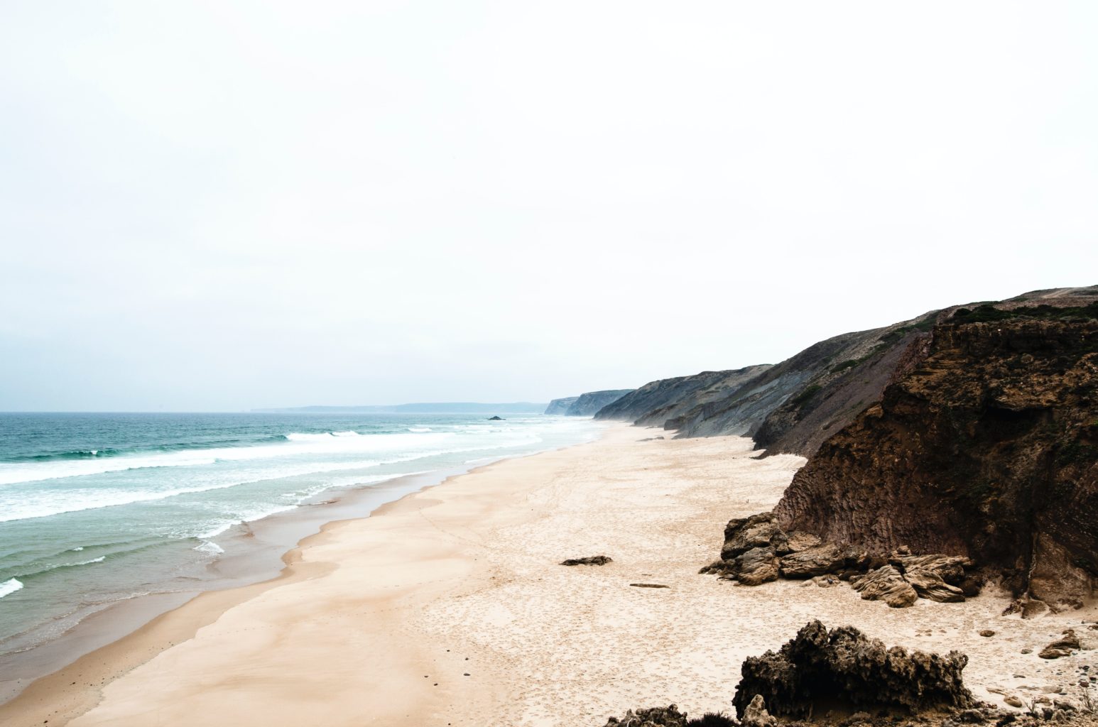 Ein leerer Strand, der von steilen Felsklippen begrenzt wird. Der Praia da Bordeira. Foto: Unsplash/Jojo Franke.